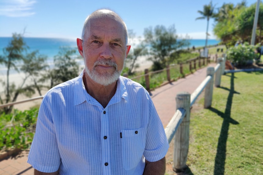 Chris Mitchell, wearing a white collared shirt with a grey beard, standing in footpath in front of beach