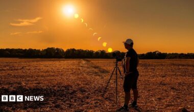 A person stands beside a camera on a tripod in a harvested field in Caterham, Surrey, during sunset. A sequence of the eclipse is shown across the sky, tracking the Sun as the Moon moves across it from high above the horizon to near sunset. Trees line the horizon beneath an orange sky.