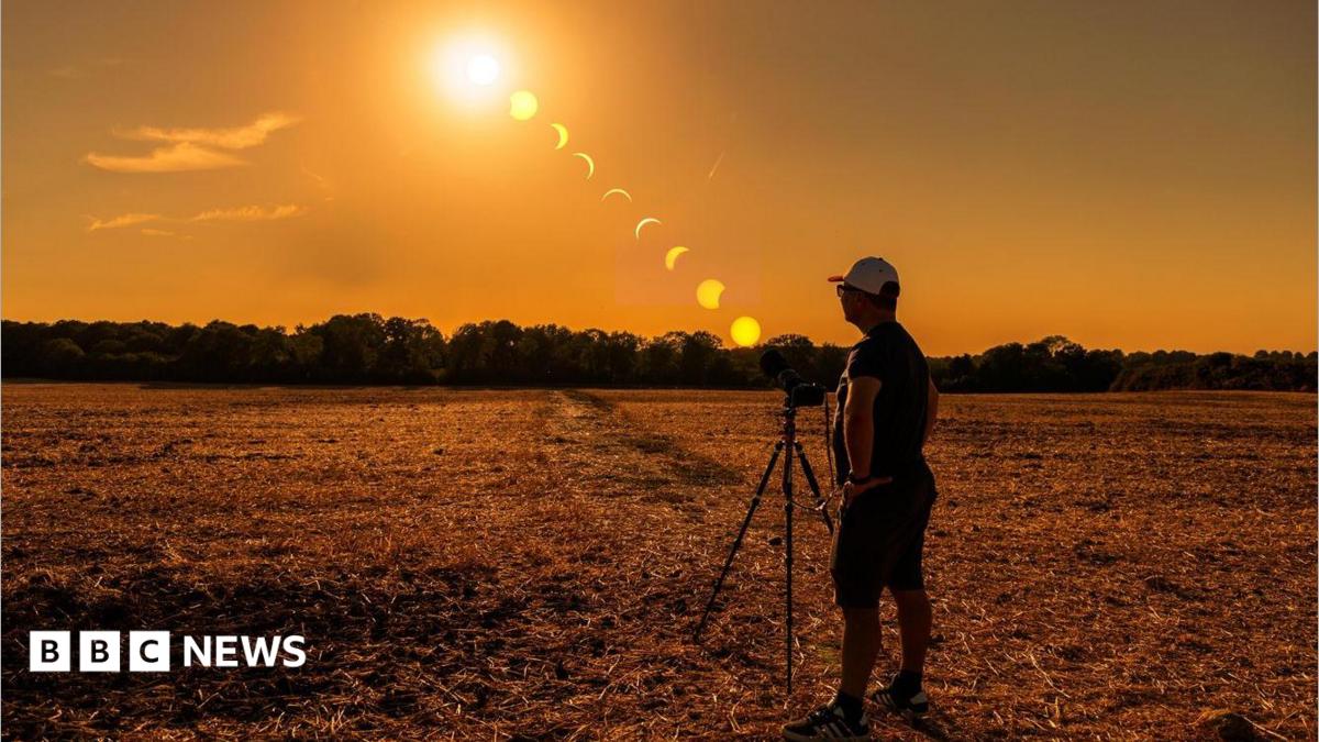 A person stands beside a camera on a tripod in a harvested field in Caterham, Surrey, during sunset. A sequence of the eclipse is shown across the sky, tracking the Sun as the Moon moves across it from high above the horizon to near sunset. Trees line the horizon beneath an orange sky.