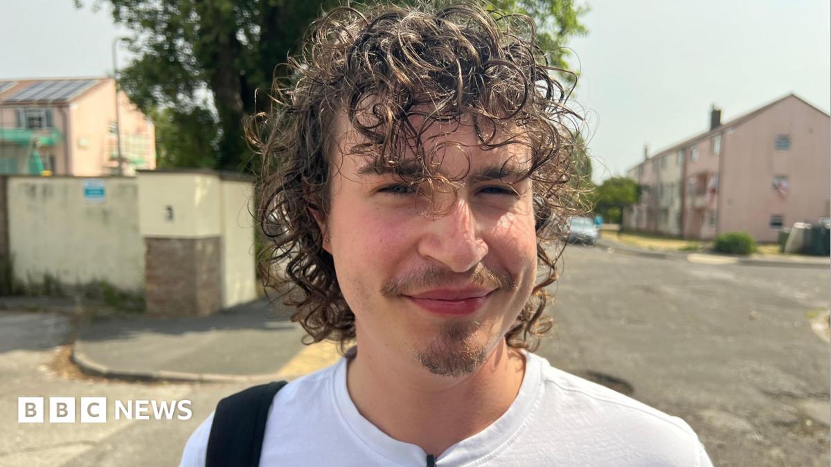 Archie Byron is smiling at the camera. the image is a head and shoulders shot of Archie. He has dark curly hair and a small beard on his chin. He is wearing a white T-shirt and has a bag over one shoulder. He is stood in an urban area with a block of flats in the background.
