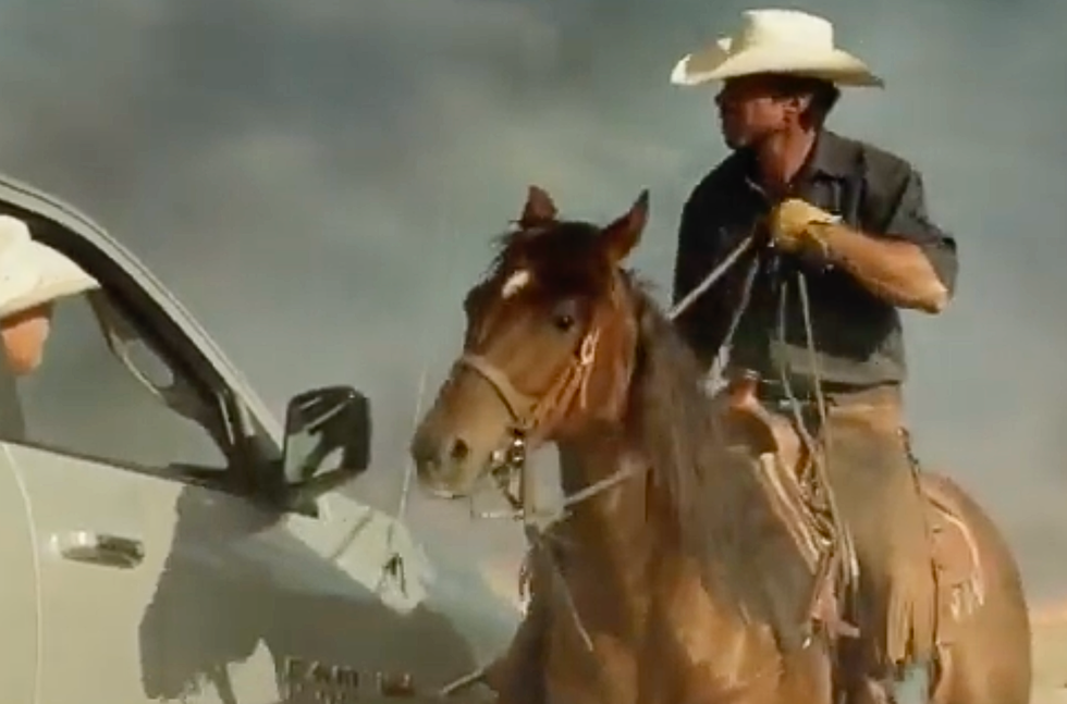 A cowboy on horseback communicates with a person in a white vehicle against a cloudy sky.