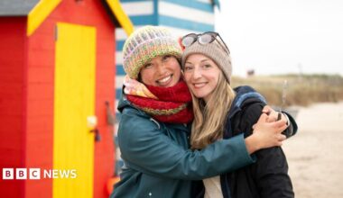 Two people stood on a beach in the winter. The woman on the right is wearing a big green coat and wooly hat. The woman on the right had a grey wooly hat and a dark coat. They are hugging, stood in front of a beach hut.