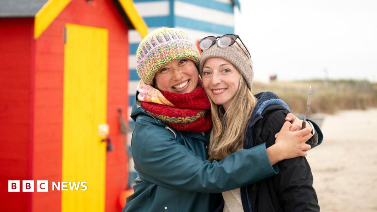Two people stood on a beach in the winter. The woman on the right is wearing a big green coat and wooly hat. The woman on the right had a grey wooly hat and a dark coat. They are hugging, stood in front of a beach hut.