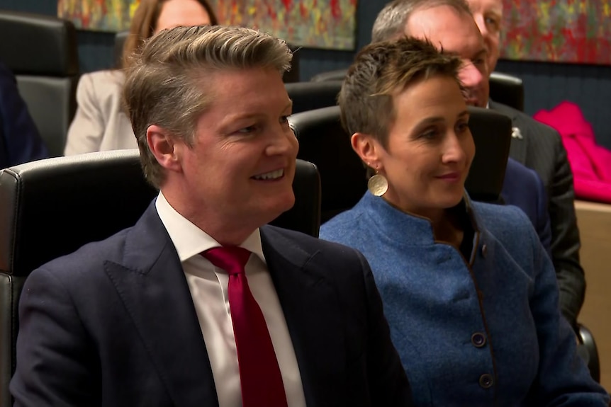 A sandy haired man in a navy suit and a dark haired woman in a blue coat sit in black chairs, smiling.