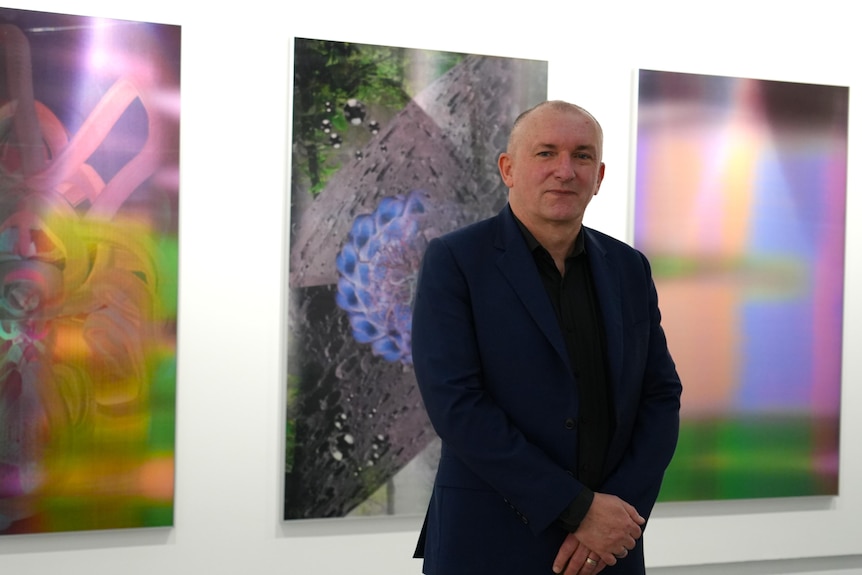 A man stands in front of three bright photographs hanging on a white wall.