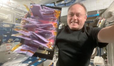 a man in a black t-shirt holds up a tray of plastic pouches filled with purple fermented milk aboard a space station.