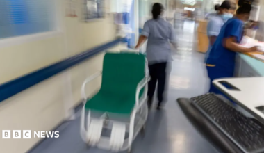 A nurse is moving a hospital chair along a corridor while two other nurses attend to paperwork