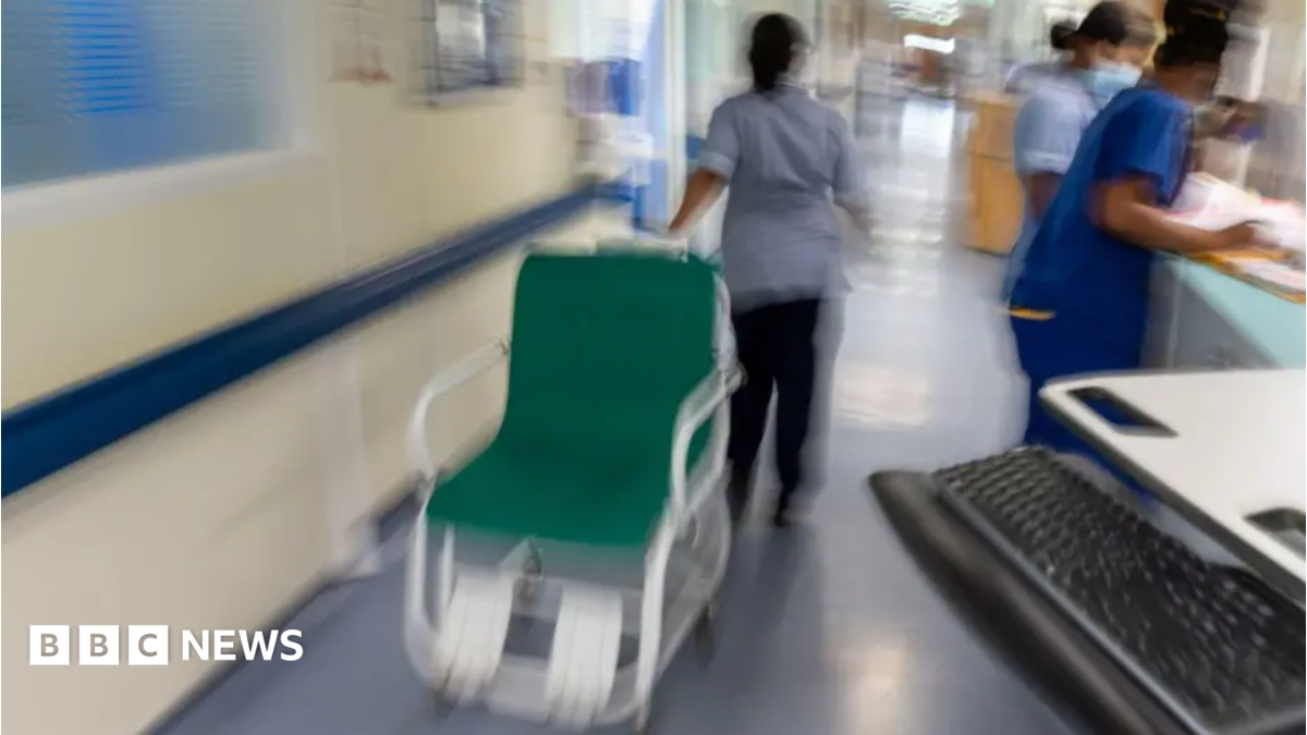 A nurse is moving a hospital chair along a corridor while two other nurses attend to paperwork