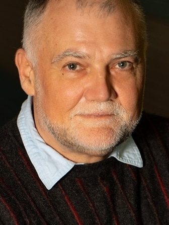 A middle-aged man with a light beard wears a dark jumper as poses for a headshot against a dark backdrop.