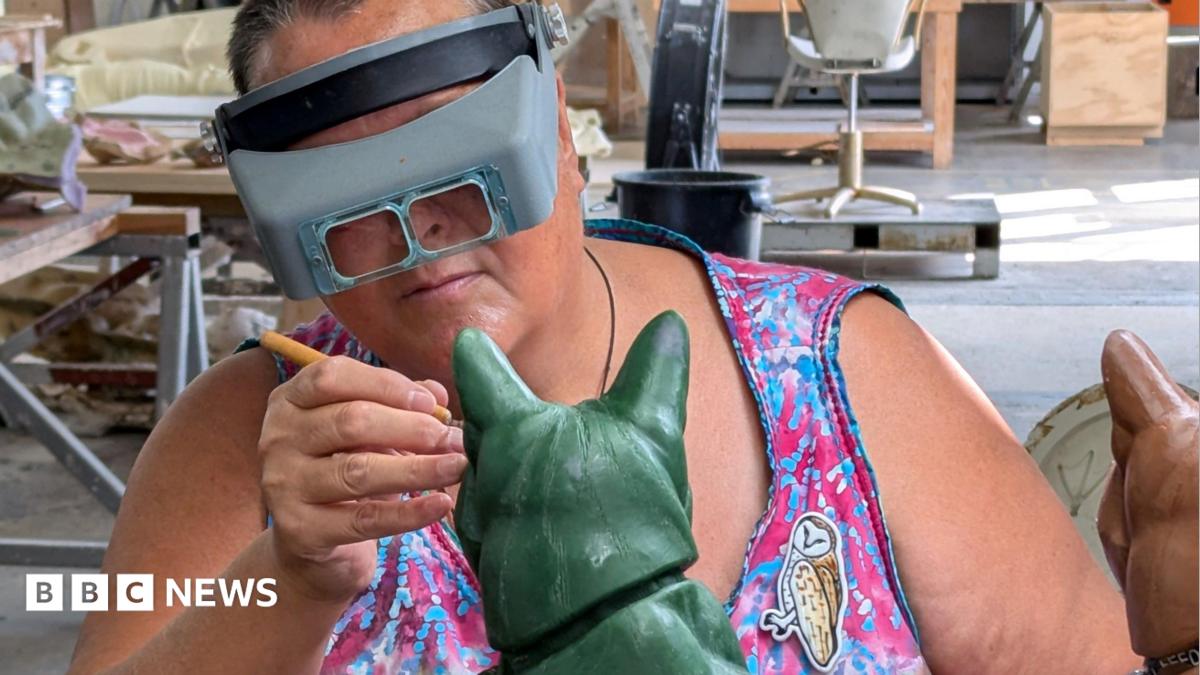 Ama Menec works on the green wax cast of Susie the cat. She is wearing grey goggles and is wearing a sleeveless pink and blue tie-dye dress. To her left is the terracotta-coloured plastolin sculpture.