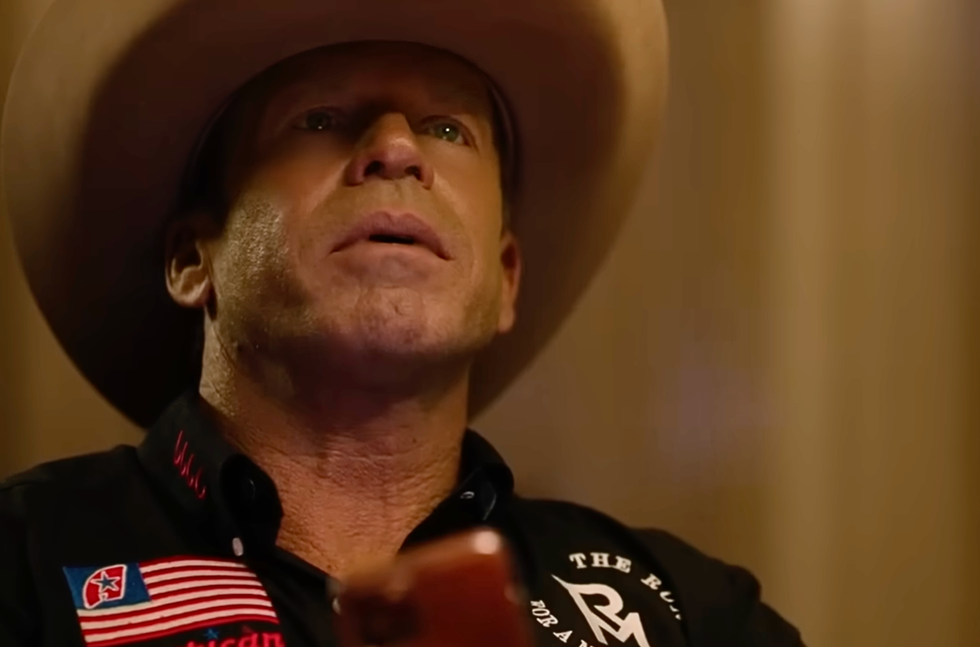 A man in a cowboy hat looks up thoughtfully while holding a phone.
