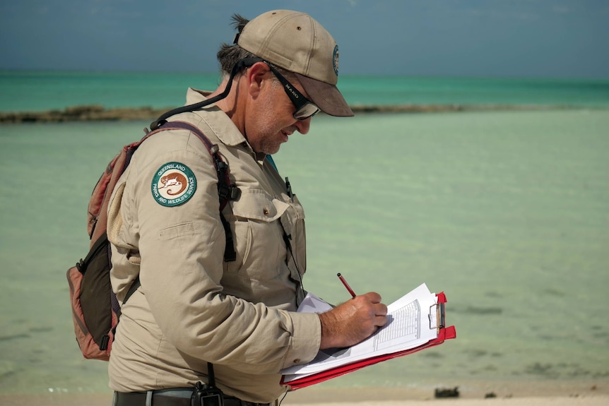 A man in a khaki uniform writes on a clipboard while standing in front of the ocean.