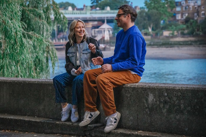 Nicola Walker and Jemaine Clement in Alice and Steve, sat on a wall by a river laughing together.