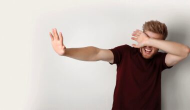 A man with reddish-brown hair and a beard wearing a maroon t-shirt holds one hand out in a stop gesture while covering his eyes with his other hand.