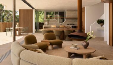 An earthy living room with a curved sectional sofa, brown suede chairs, a wood coffee table, a vase of tulips, a brown hanging fireplace, a stone kitchen, and the balcony doors drawn open