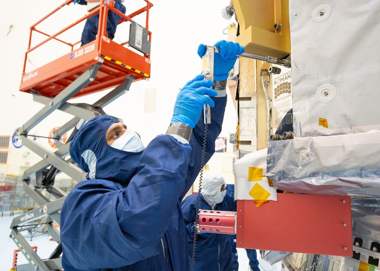 People in blue protective suits and a red cherry picker work in an industrial high bay.