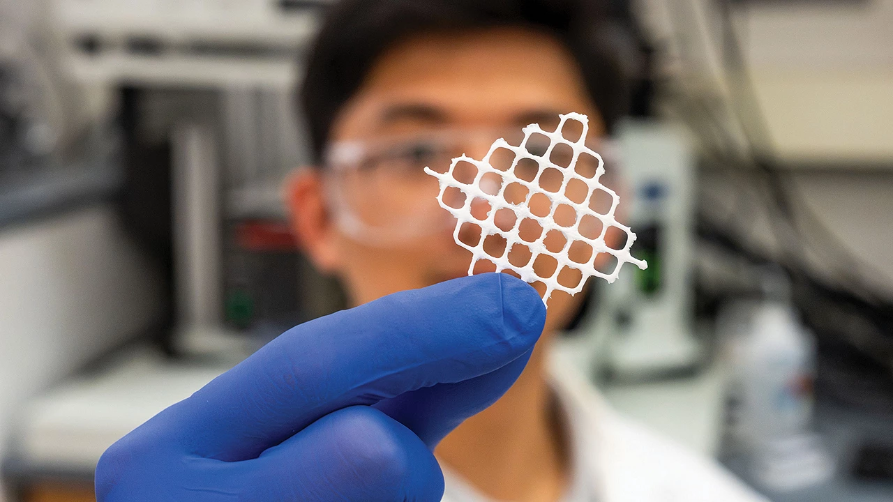 A researcher, blurry in the background, holds a square of white mesh in a gloved hand.