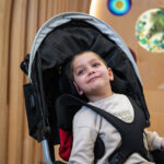 Child secured in a stroller with a padded harness, seated indoors in front of a wood wall with colorful circular decorations.