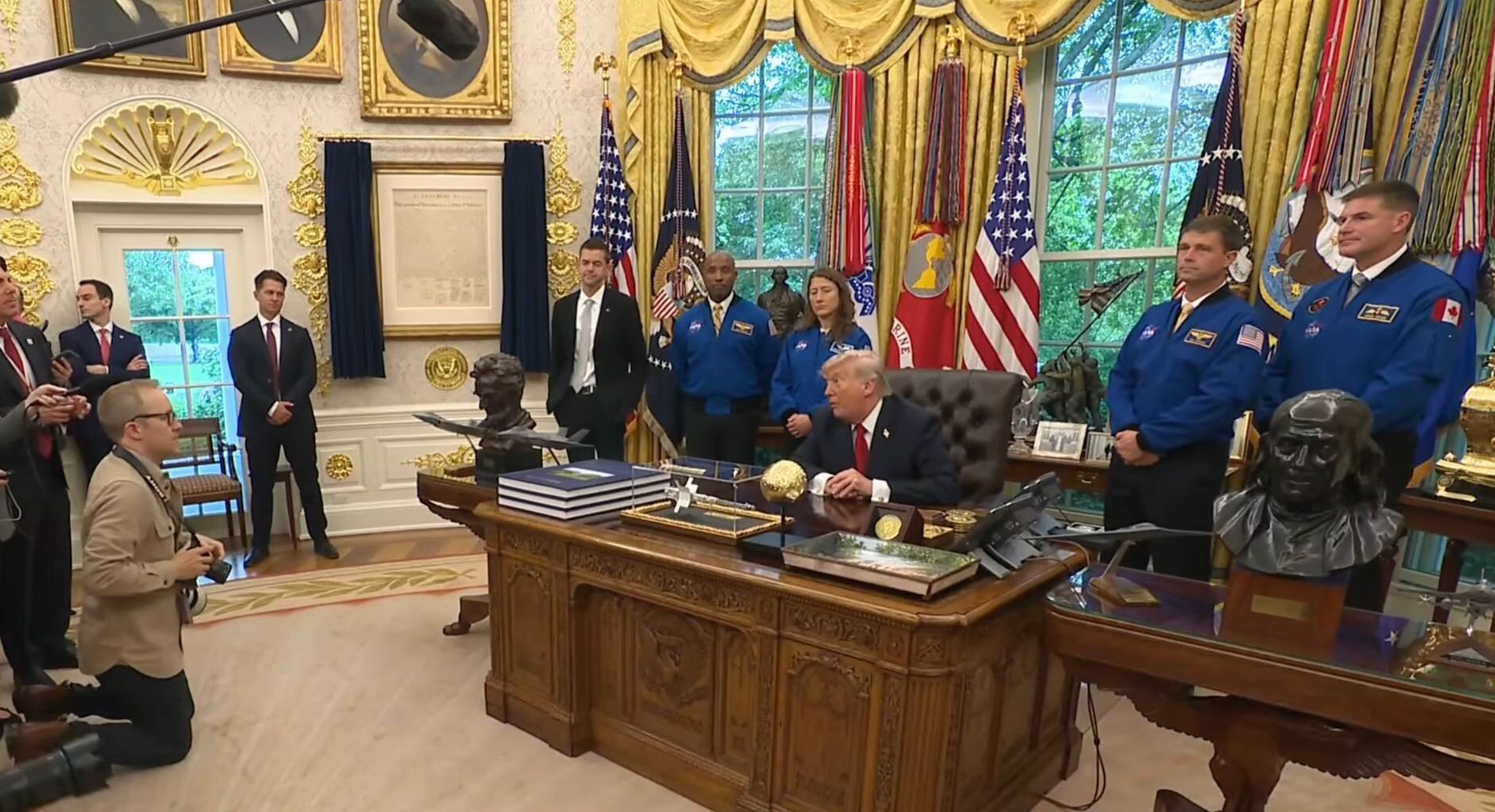 four people in blue flights stand behind a man in a suit sitting at a fancy desk