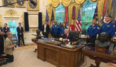 four people in blue flights stand behind a man in a suit sitting at a fancy desk
