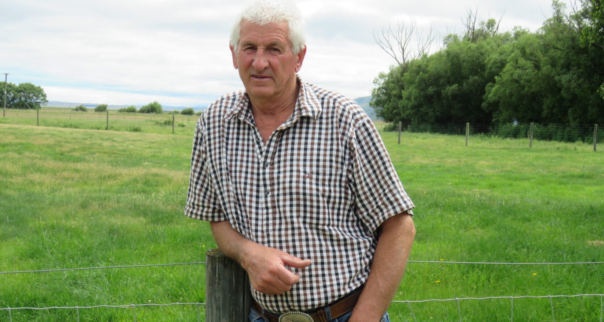 Older man wearing a plaid shirt leans on a wooden fence post in a grassy rural field with trees in the background