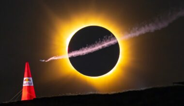 A small aircraft with a smoke trail flies in front of a solar eclipse, positioned above a red Red Bull branded pylon on a dark horizon.
