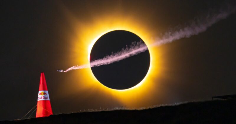 A small aircraft with a smoke trail flies in front of a solar eclipse, positioned above a red Red Bull branded pylon on a dark horizon.