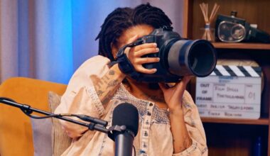 A person with dreadlocks holds a large camera up to their face, partially obscuring it, while sitting in front of a microphone and a film clapperboard on a shelf.