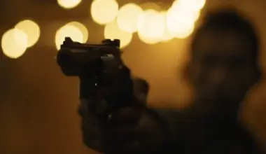 A person is pointing a handgun toward the camera in a dimly lit setting with blurred lights in the background.