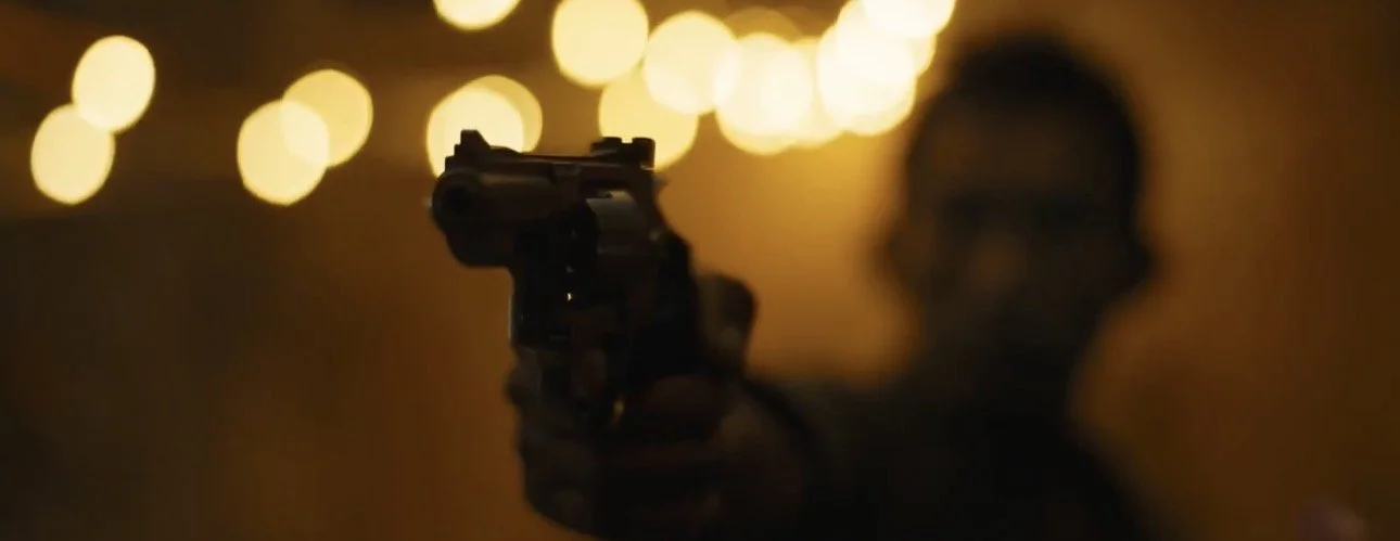 A person is pointing a handgun toward the camera in a dimly lit setting with blurred lights in the background.