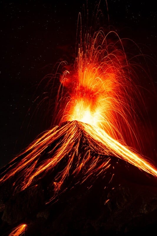 A volcanic eruption at night, with glowing lava spraying into the sky and flowing down the dark, conical mountainside.