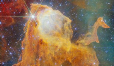 A vibrant nebula of orange and yellow gas clouds illuminated by bright, star-like points of light with distinct diffraction spikes.