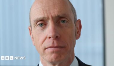 A bald man with blue eyes looking directly at the camera. Behind him is white blinds and a cream wall.