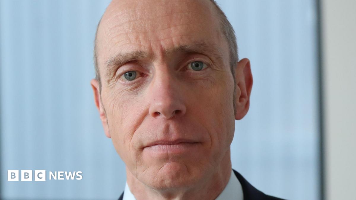 A bald man with blue eyes looking directly at the camera. Behind him is white blinds and a cream wall.