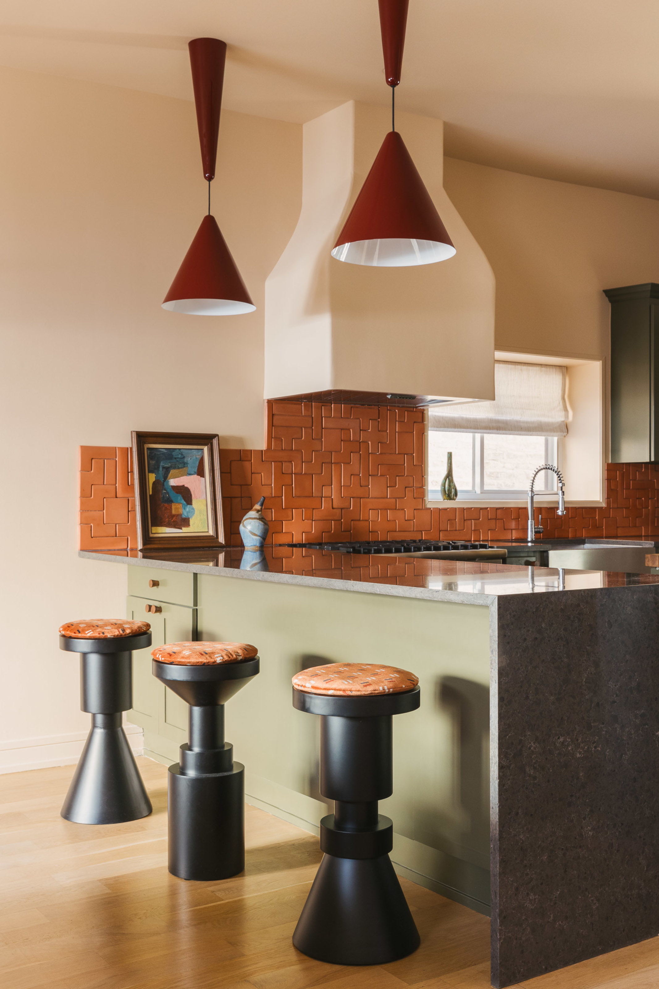 U-shaped kitchen with sage green units, grey stone waterfall countertop, terracotta geometric backsplash, black sculptural bar stools and red pendant lights