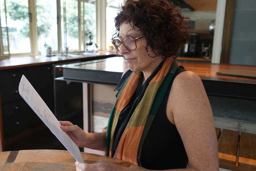 A woman wearing glasses and a black sleeveless top sits looking at a piece of paper with writing on it. Kitchen behind her.