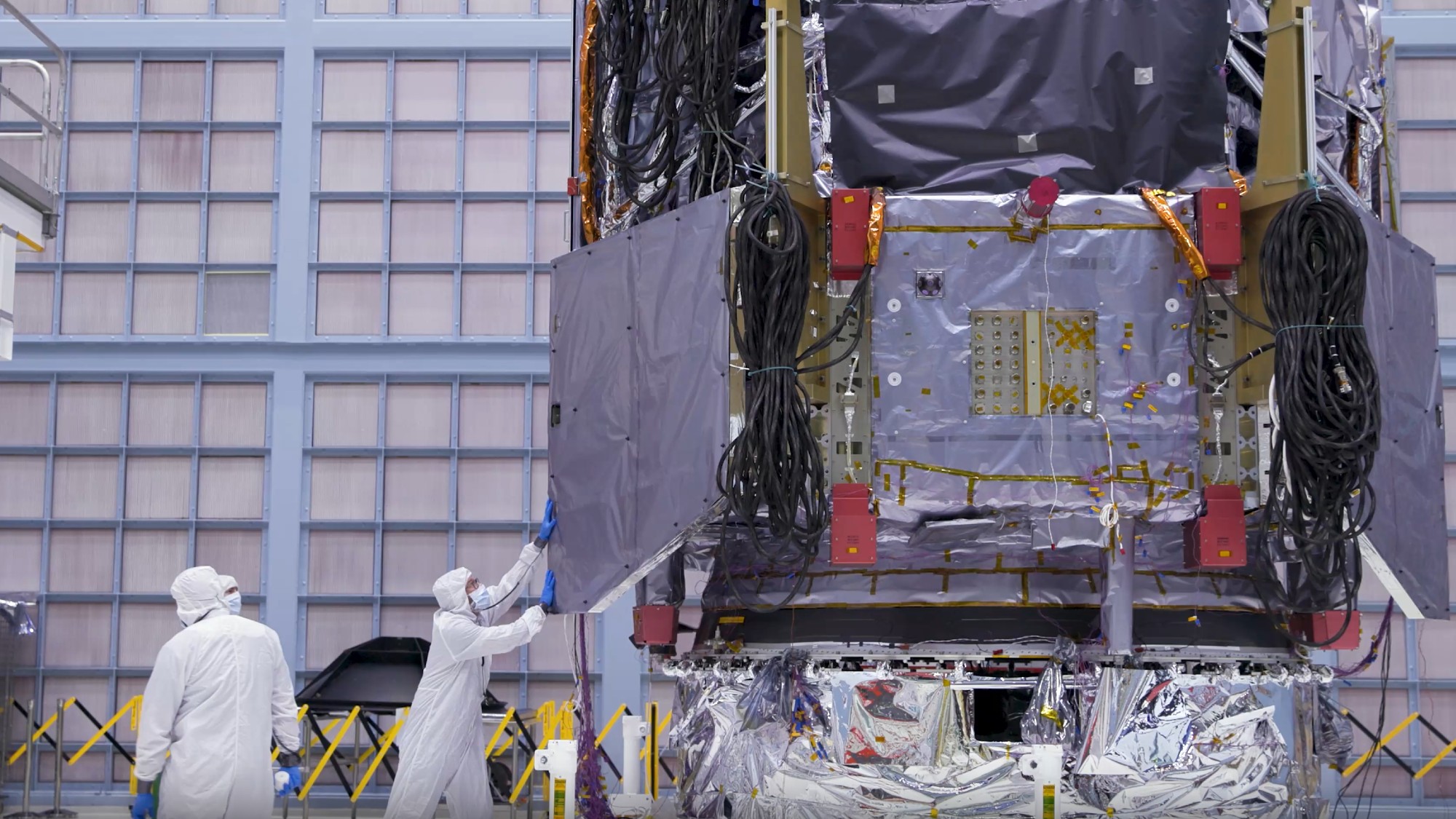 technicians in clean suits in a hangar next to a large rectangular spacecraft wrapped in metallic foil