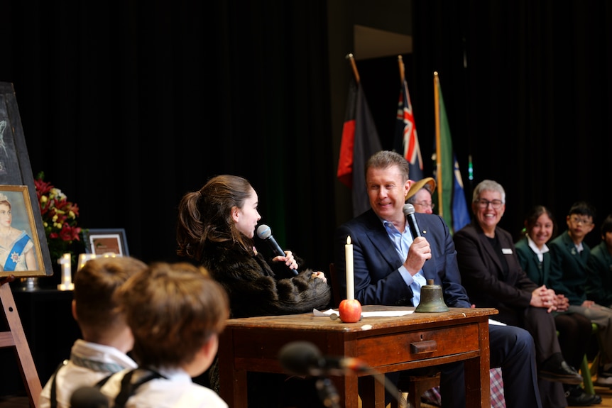 A man wearing a suit on stage holding a microphone, a student doing the same, others looking on