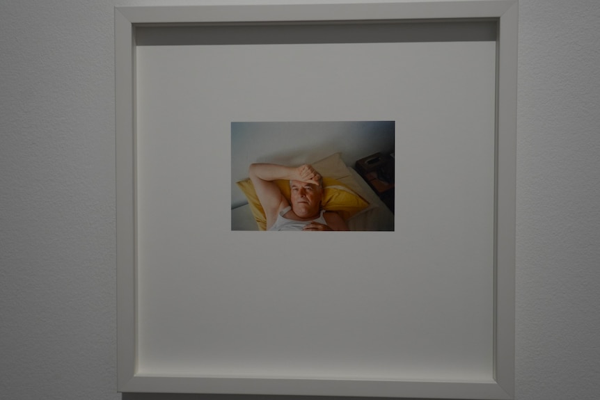 Small photograph of man lying on a pillow on a bed with his hand on his forehead in a large white frame.
