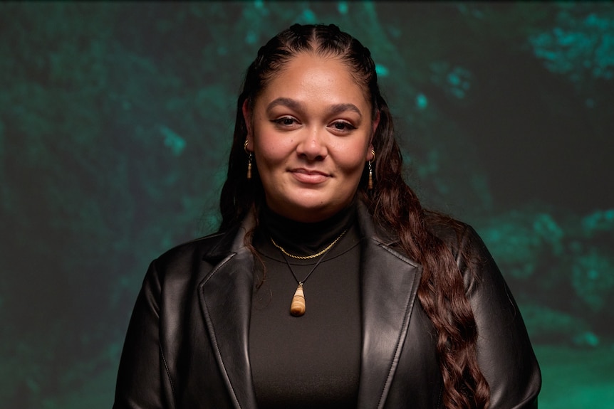 A woman with dark hair and tan skin stands in front of a green background.