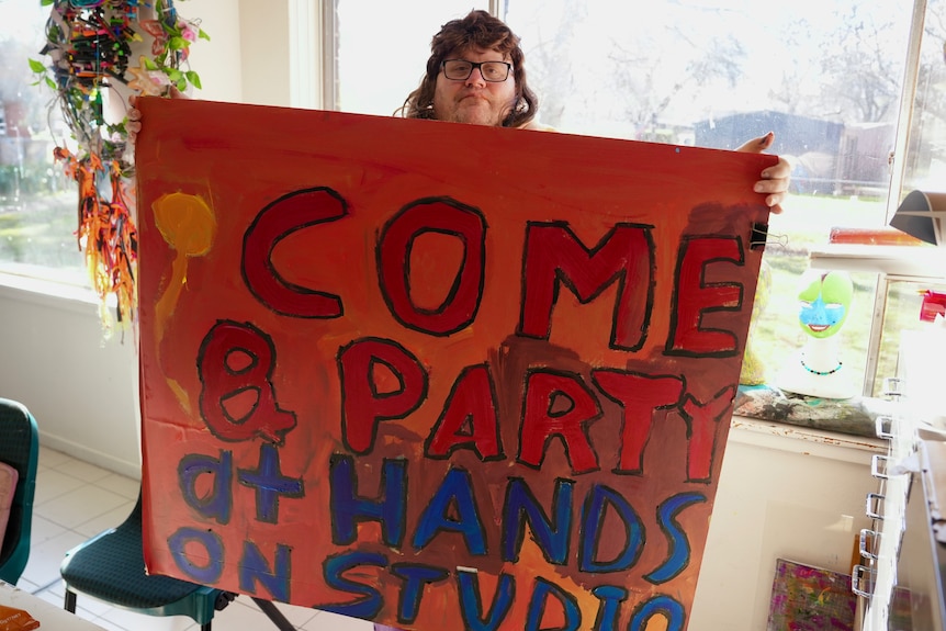 A man holds up a large artwork with the words "Come and party at Hands on Studio" painted on it.
