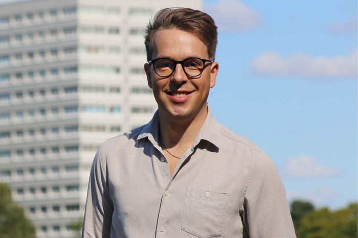 Ben Wadey stands in front of a tower block. He is wearing a grey shirt and glasses and is smiling
