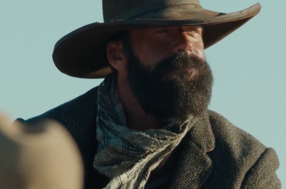 A rugged man with a hat and a beard, wearing a scarf and a jacket, looking off into the distance against a clear sky.