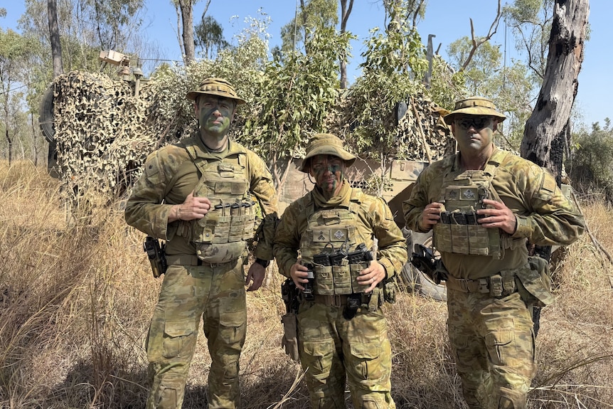 Three people stand together in army uniforms in front of scrubby bushland