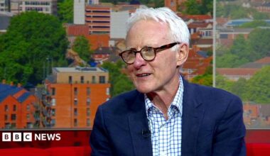 Norman Lamb sitting on the red sofa in the BBC Look East studio. He is looking off to the left in the direction of the presenter who cannot be seen in the picture. He is wearing a white and blue checked shirt with a navy blazer over the top.