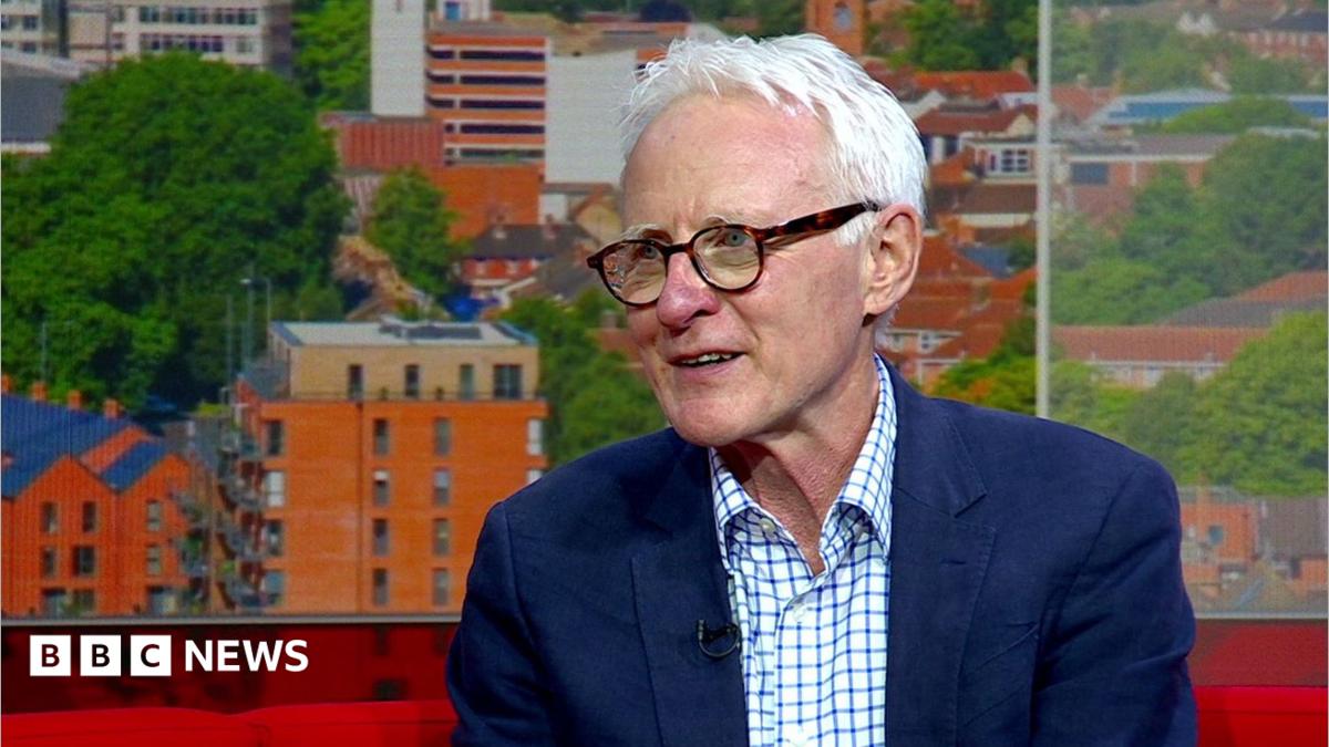 Norman Lamb sitting on the red sofa in the BBC Look East studio. He is looking off to the left in the direction of the presenter who cannot be seen in the picture. He is wearing a white and blue checked shirt with a navy blazer over the top.