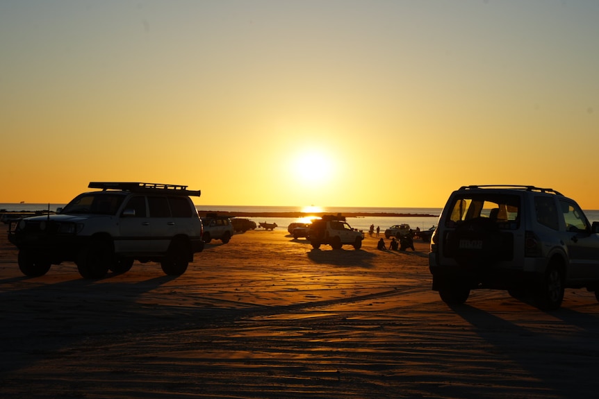 Four-wheel drives on a beach bathed in yellow light as the sun sets over the water.
