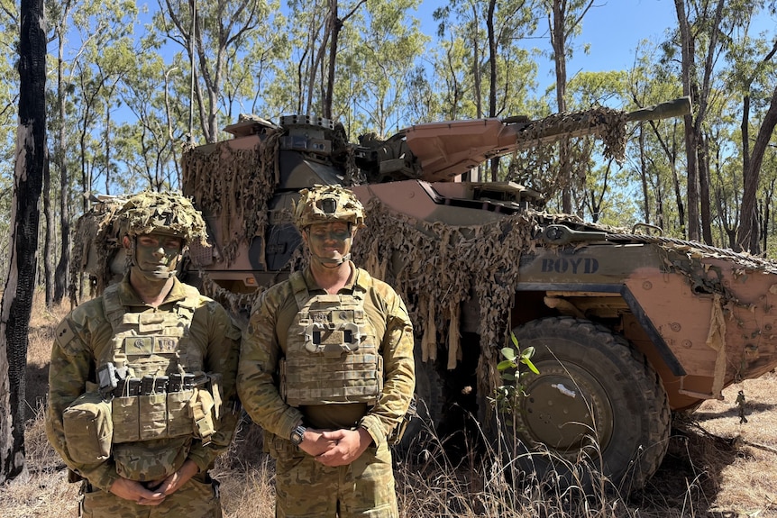 Two men standing in front of a large military vehicle in scrubby bushland