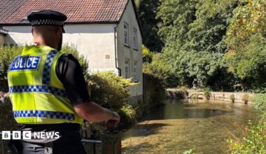 A police officer walking on a raised footpath alongside a shallow, clear river. On the opposite side is a white rendered cottage with a chimney and red tiled roof.