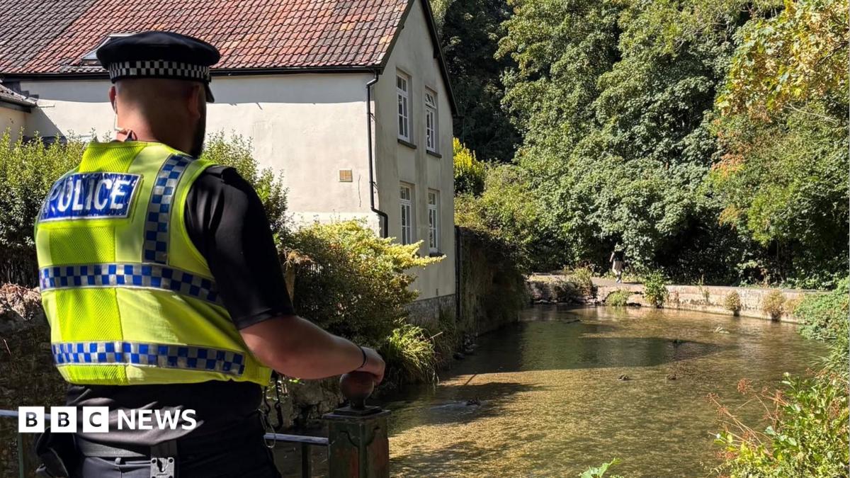 A police officer walking on a raised footpath alongside a shallow, clear river. On the opposite side is a white rendered cottage with a chimney and red tiled roof.